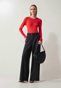 Red long-sleeve top with a fitted design, paired with high-waisted black wide-leg trousers. The model holds a black hobo bag.