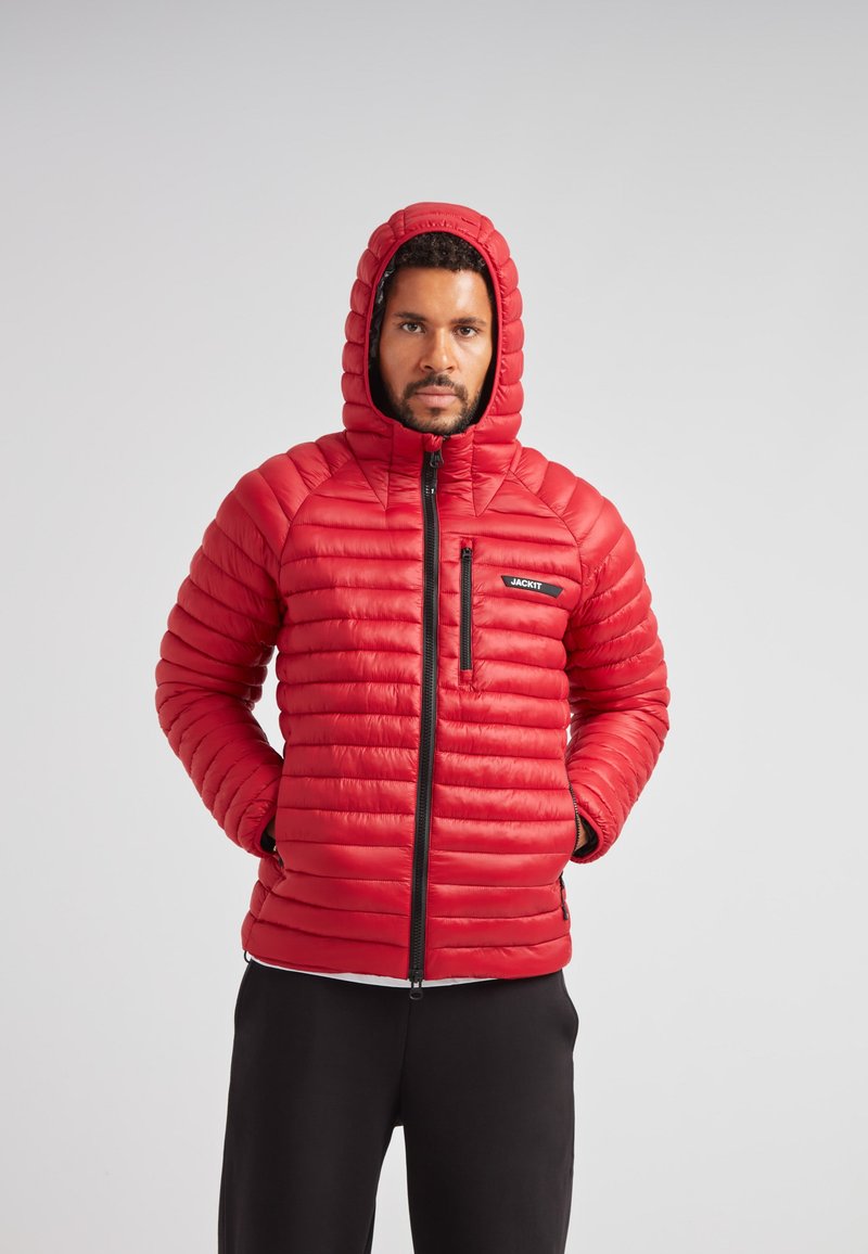 Giacca piumino rossa con apertura a zip, cappuccio e texture a coste. Presenta una tasca sul petto con un logo nero e polsini elastici.
