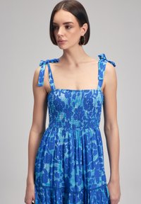Abito blu con corpino arricciato e motivo floreale. Presenta spalline regolabili e gonna a strati. Tessuto morbido e testurizzato.