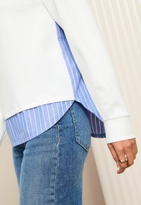 Sudadera blanca con una capa inferior a rayas azules, que presenta una abertura lateral. Combinada con jeans de mezclilla azules. Primer plano de la manga y la mano con un anillo.