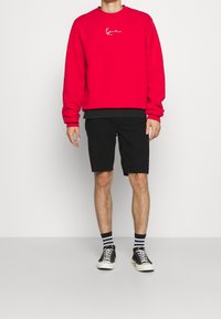 Felpa rossa con colletto rotondo e dettaglio del logo bianco. Pantaloni shorts neri con orlo sfrangiato e scarpe da ginnastica nere con suola bianca e calzini a righe.