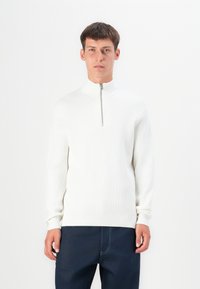 Suéter blanco de canalé con cuello alto y cierre de media cremallera. Presenta mangas largas y una silueta ajustada. Se lleva con pantalones oscuros.