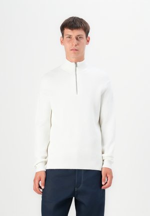 Suéter blanco de canalé con cuello alto y cierre de media cremallera. Presenta mangas largas y una silueta ajustada. Se lleva con pantalones oscuros.