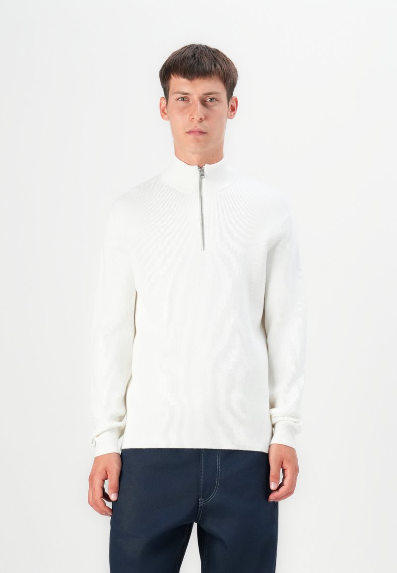 Suéter blanco de canalé con cuello alto y cierre de media cremallera. Presenta mangas largas y una silueta ajustada. Se lleva con pantalones oscuros.
