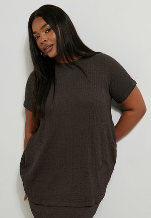 Donna con lunghi capelli scuri che indossa una maglia a maniche corte marrone scuro testurizzata, in piedi davanti a uno sfondo grigio chiaro.