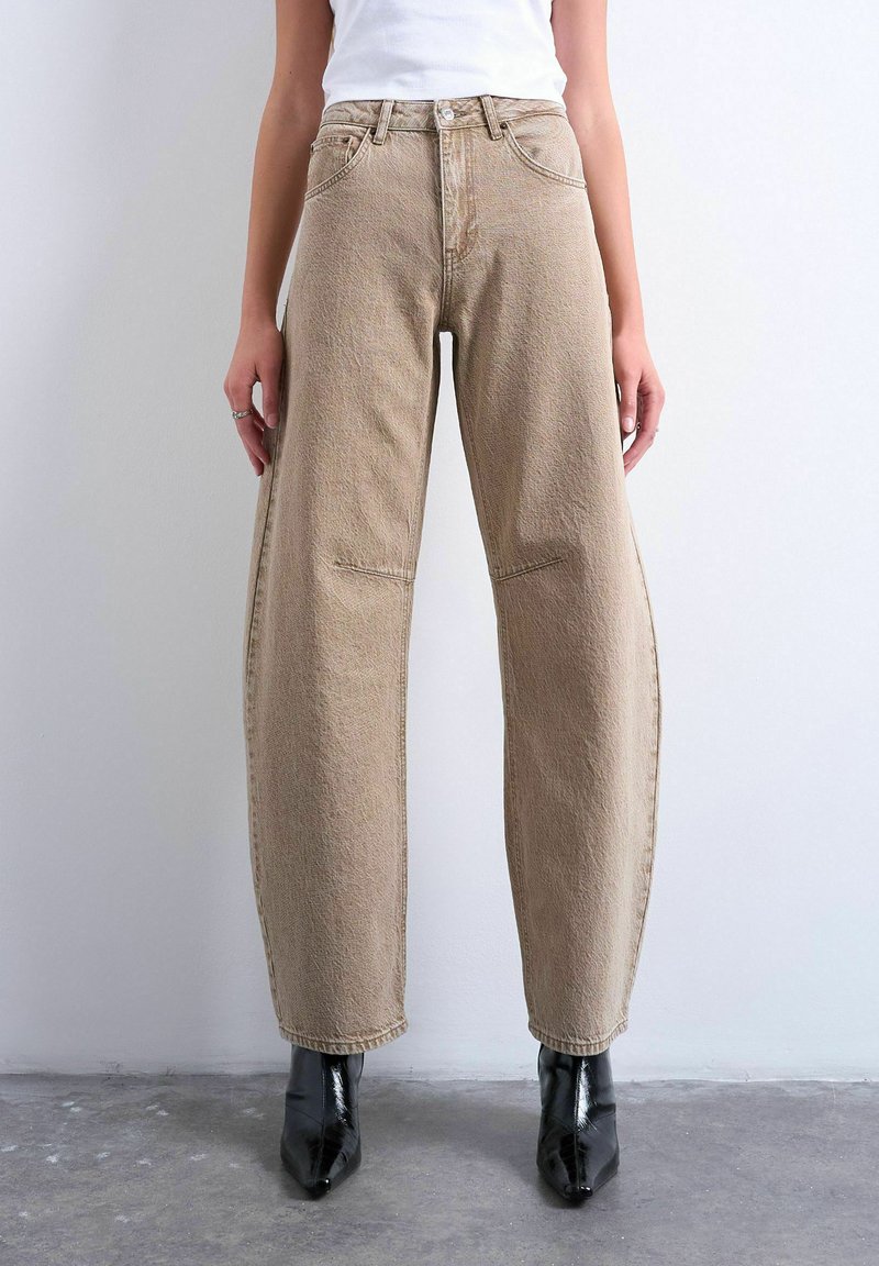 Kvinde iført beige jeans med høj talje og løs pasform, kombineret med sorte spidse støvler, stående op ad en ensfarvet lys væg.