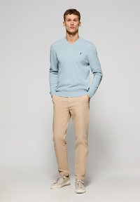 Jasnoniebieski sweter z dekoltem w serek i prążkowanymi mankietami, w połączeniu z beżowymi spodniami i jasnymi trampkami. Subtelne logo na piersi swetra.