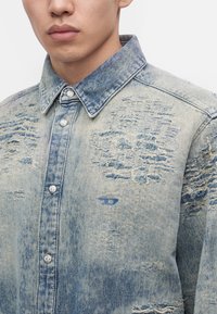 Jeune homme portant une veste en jean bleu clair usée avec des boutons-pression argentés et des patchs effilochés sur la poitrine et l’épaule.
