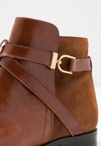 Brune ankelstøvle med en blanding af glat læder og nubuck, med en guldtonet spænde og remdetalje samt en tekstureret overflade.