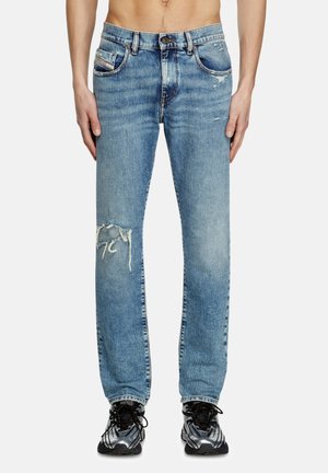 Mann trägt hellblaue verwaschene Jeans mit einem Riss am linken Knie und schwarze sowie silberne Sportschuhe.