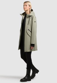 Naine seismas helerohelises kapuutsiga vihmajopes, millel on mustad tõmblukud, mustad retuusid ja mustad poolpõlve saapad tavalise tausta ees.