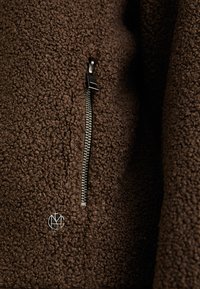 Close-up of a brown textured fleece fabric with a vertical silver zipper and a small metallic circular logo below it.