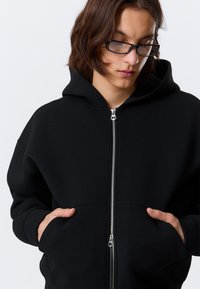 Felpa nera con zip, dotata di cappuccio, realizzata in un tessuto morbido. Presenta tasche frontali e chiusura con zip argentata. Design semplice e monocromatico.