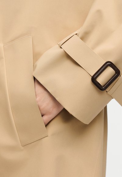 Χέρι στην τσέπη του μανικιού μιας μπεζ καμπαρντίνας, που διαθέτει ταιριαστή λωρίδα με σκοτεινή ορθογώνια αγκράφα.