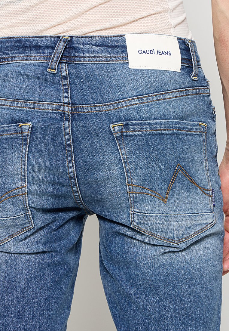 Widok z tyłu osoby noszącej niebieskie dżinsy Gaudí z przeszytymi tylnymi kieszeniami i białą siateczkową bluzkę.