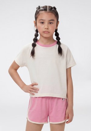 Fată tânără cu păr împletit stă cu o mână pe șold, purtând un tricou alb cu margini roz și pantaloni scurți roz cu margini albe.
