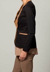 Blazer noir avec col et bordure beige, présentant un design structuré, un style à un seul bouton et des poches latérales. Silhouette simple et ajustée.
