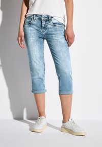 Jean capri en denim bleu clair avec des poignets retroussés, comportant cinq poches et de subtils accents usés. Associé à des baskets blanches.