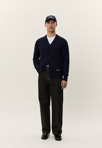 Άνδρας που φοράει μπλε σκούρη ζακέτα, λευκό t-shirt, μαύρο φαρδύ παντελόνι, μαύρα παπούτσια και μπλε σκούφο baseball, στέκεται μπροστά σε απλό φόντο.