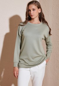 Top de manga larga verde claro con un ajuste relajado, puños acanalados y una textura suave, combinado con pantalones blancos. El fondo es neutral.