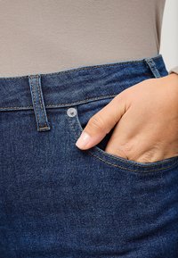 Donkerblauwe jeans met een klassieke pasvorm, voorzien van een voorzak, knoopsluiting en contrasterende stikseldetails. Zachte denimtextuur is zichtbaar.