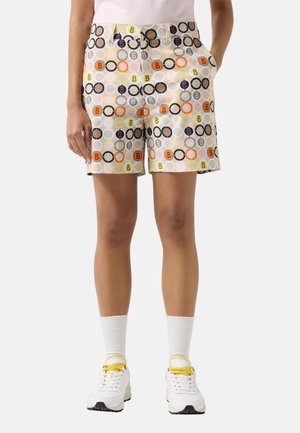 Vrouw draagt kleurrijke shorts met geometrisch patroon, witte sokken, witte sneakers met gele veters en een lichtroze top, staand met handen in de zakken.