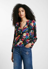 Schwarze Bluse mit langen Ärmeln und Blumenmuster, verziert mit lebhaften roten, blauen und gelben Blumen. Sie hat eine lockere Passform und einen V-Ausschnitt. Kombiniert mit Jeans.