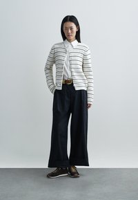 Cardigan blanc en tricot avec des rayures horizontales, associé à une chemise blanche, un pantalon large noir et des chaussures marron à lacets. Ceinture dorée en accent.