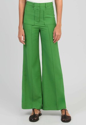 Pantalon large taille haute vert avec grandes poches à rabat sur le devant, porté avec des sandales plates tressées noires.