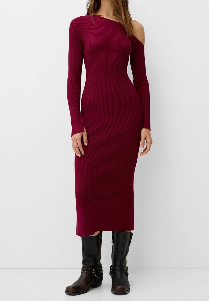 Femme portant une robe midi bordeaux ajustée en maille côtelée à manches longues avec une épaule dégagée, associée à des bottes noires mi-mollet avec des sangles.