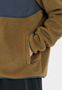 Brun fleece-pullover med en marinblå bröstpanel, med en mjuk textur, sidofickor och elastiska ärmslut för ökad värme.