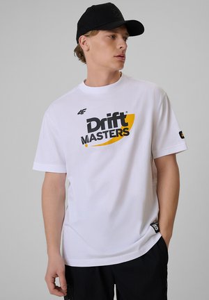 Młody mężczyzna w białej koszulce "Drift Masters" i czarnej czapce, patrzy w lewo z jedną ręką w kieszeni.