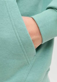 Miętowa zielona bluza z miękką fakturą, z przednią kieszenią i ściągaczami na mankietach. Zbliżenie podkreśla szczegóły przeszyć.