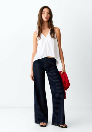 Mujer con una camiseta blanca sin mangas, pantalones anchos de raya diplomática azul marino, sandalias negras, sosteniendo una chaqueta roja en la mano izquierda.