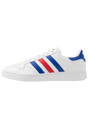 Baskets basses blanches Adidas avec des bandes bleues et rouges sur le côté, semelle blanche et languette de talon bleue avec logo.