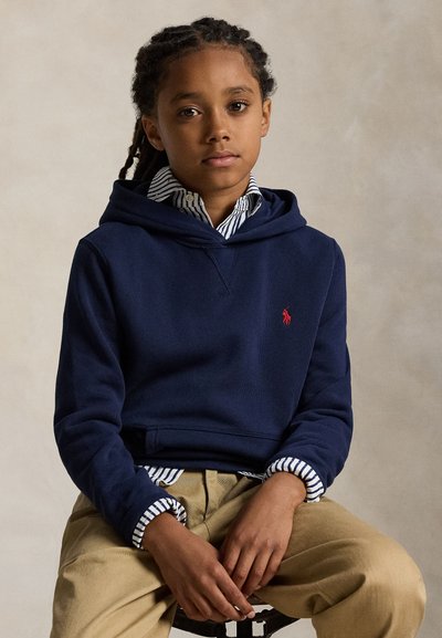 Vaikas su pintomis kasomis sėdi dėvėdamas tamsiai mėlynį gobtinį per dryžuotą marškinius ir smėlio spalvos kelnes neutralaus fono akivaizdoje.