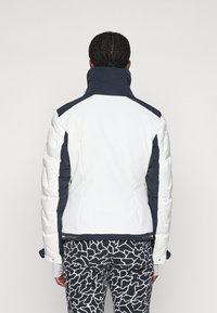 Chaqueta acolchada blanca y azul marino con cuello alto, que presenta un diseño elegante y mangas texturizadas. Combinada con pantalones negros de estampado.