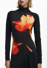 Frau in einem schwarzen langärmeligen Kleid mit einem verschwommenen, leuchtenden rot-orangefarbenen Blumenmuster auf dem Oberteil.