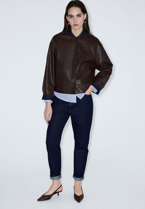 Mujer con chaqueta de cuero marrón oscuro, jeans azul marino remangados, camisa de rayas y zapatos slingback marrones de tacón bajo, posando frente a un fondo blanco.