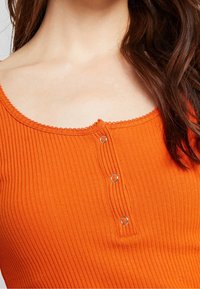 Top naranja acanalado con escote amplio, vuelta decorativa en forma de concha y tres botones metálicos en la parte frontal. Tela suave y elástica.