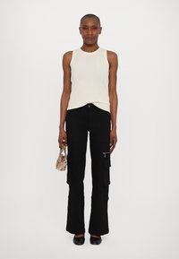 Mujer vestida con un top de punto blanco sin mangas y pantalones cargo negros, sosteniendo un pequeño bolso estampado, de pie contra un fondo blanco sencillo.