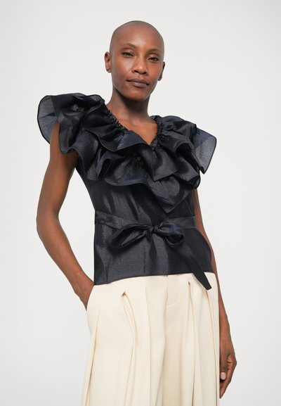 Sort tekstureret top med store flæseærmer og V-hals, med en bundet talje-bånd. Sammen med cremefarvede plisserede bukser.