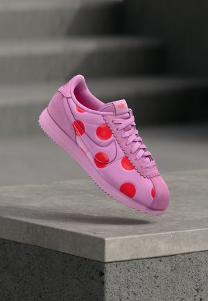 Scarpe da ginnastica rosa con pois rossi e suola texturizzata, sospese a mezz'aria sopra una sporgenza di cemento con scale sfocate sullo sfondo.