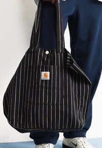 Bolsa de tela de rayas en blanco y negro con un bolsillo frontal y un parche con logo, que cuenta con asas dobles para llevar.