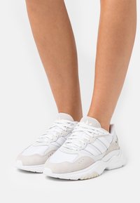 Vita idrottssneakers i vitt med grå mockadetaljer, snörning och framträdande tre ränder på sidan. Texturerad gummisula.