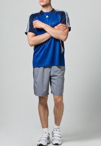 Modré sportovní tričko s krátkým rukávem a námořnickými akcenty, kombinované se šedými šortkami. Obsahuje tři bílé pruhy na ramenou a bílé ponožky se sandály.