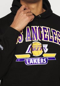 Felpa nera con la scritta "LOS ANGELES LAKERS" in grassetto giallo e viola. Presenta una grafica di un pallone da basket e dettagli con cappuccio regolabile.