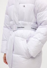 Svetlofialová prešívaná bunda s opaskom so sťahovacím efektom a logom "Calvin Klein", bočnými vreckami a prešívanou textúrou pre izoláciu.