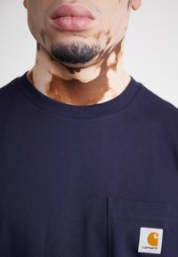 Camiseta de algodón azul marino con cuello redondo, mangas cortas y un bolsillo rectangular con el logo de Carhartt en blanco y naranja.
