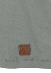 Stonowany zielony materiał z brązowym skórzanym naszywką przyszytą w pobliżu dolnego brzegu, wytłoczonym napisem i logo "KEY LARGO".